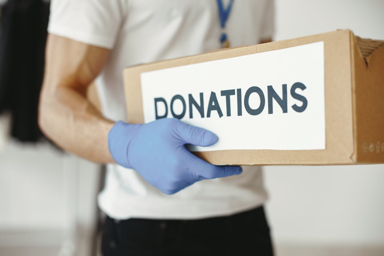 A volunteer wearing gloves holds a 'donations' box, symbolizing community support.
